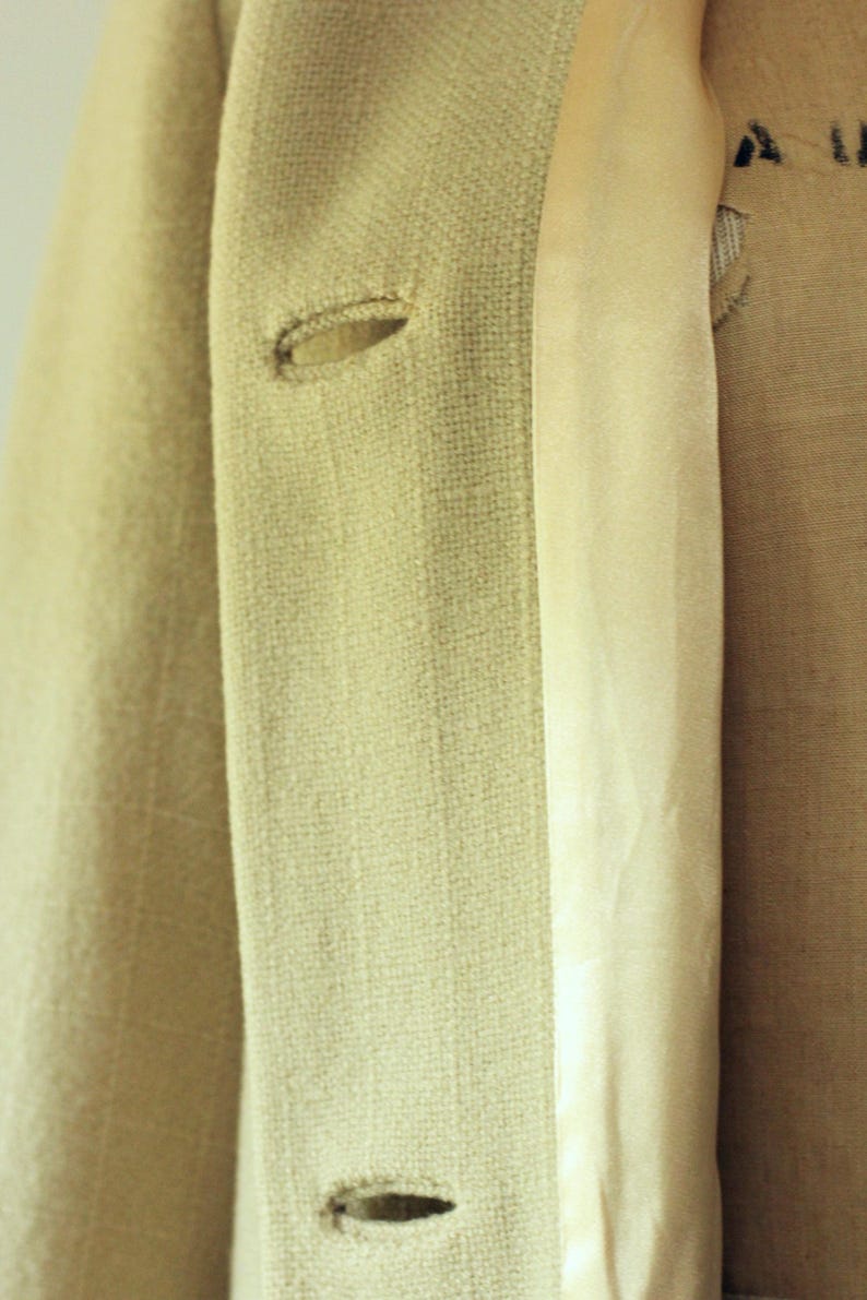 Puede incluir: Primer plano de una blazer beige claro con dos ojales visibles. La blazer tiene una tela texturizada y un forro satinado. La imagen destaca los detalles de la confecci&oacute;n.
