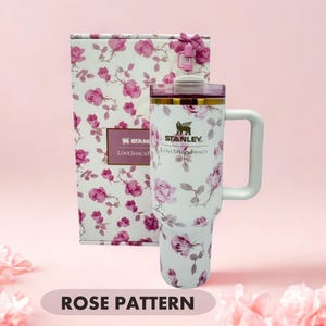 May include: A white Stanley LoveShackFancy tumbler with a pink rose pattern, a handle, and a clear lid with a pink straw topper. The tumbler is displayed in front of a matching rectangular box featuring the same floral design and brand text.