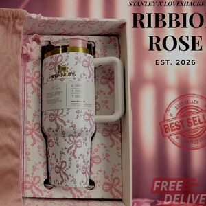Puede incluir: Un vaso Stanley blanco con un patr&oacute;n de lazos rosas, un logotipo dorado y una tapa rosa claro. El vaso se presenta en una caja a juego con una bolsa de tela rosa. El texto en la caja dice Stanley x LoveShackFancy, Ribbon Rose, Est. 2026.