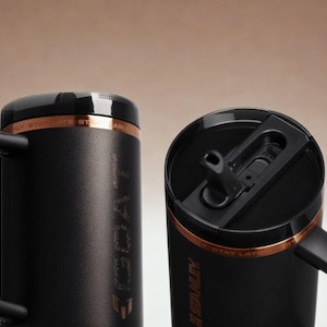 May include: Two matte black insulated tumblers with sturdy side handles and black lids. Each tumbler features a copper-coloured band near the top with the text STAY LATE. One lid shows a black straw opening, while the other is closed.