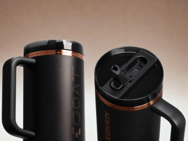 May include: Two matte black insulated tumblers with sturdy side handles and black lids. Each tumbler features a copper-coloured band near the top with the text STAY LATE. One lid shows a black straw opening, while the other is closed.