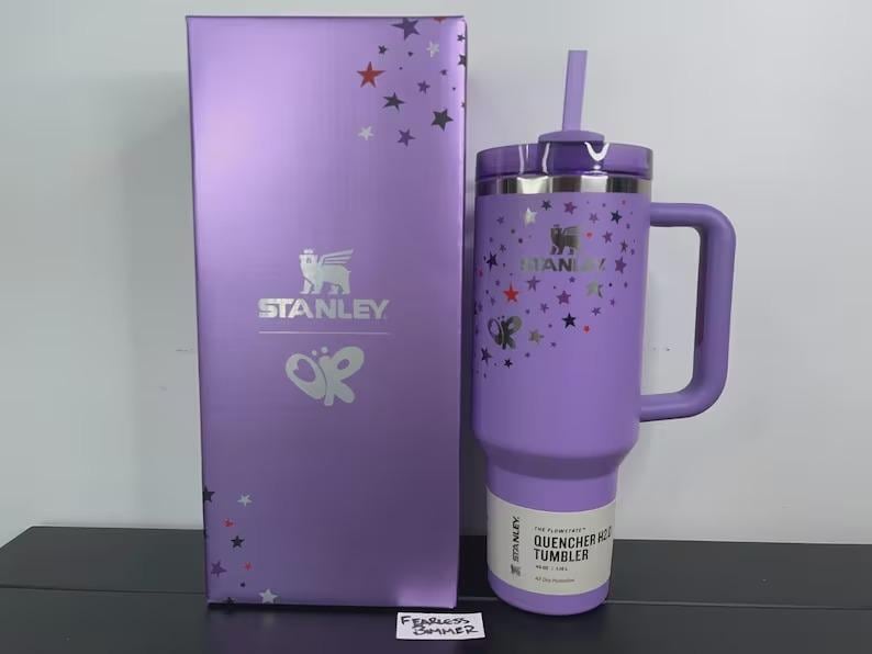 May include: A lavender Stanley Quencher H2.0 FlowState tumbler with a matching straw and handle, featuring a star pattern and OPI logo. It is displayed next to its tall, rectangular lavender box, which also displays the Stanley and OPI branding.