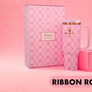 May include: A light pink Stanley tumbler with a handle and a clear lid, featuring a repeating pattern of pink ribbons. The tumbler is displayed next to a matching rectangular box with the same ribbon pattern and a Stanley LoveShackFancy label.