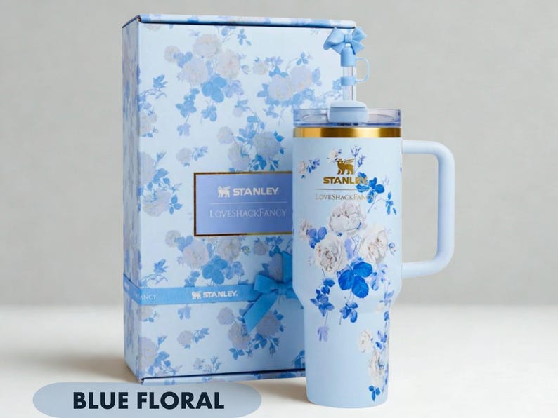May include: A light blue Stanley tumbler with a floral pattern and a matching gift box. The tumbler features a gold rim, a handle, and a straw with a small blue bow. The Stanley and LoveShackFancy logos are printed on both the tumbler and the box.