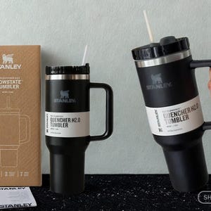 May include: Two black Stanley Flowstate Quencher H2.0 tumblers with handles, straws, and black lids. One tumbler stands next to its brown cardboard packaging, while the other is held. The packaging features the Stanley logo and product name.