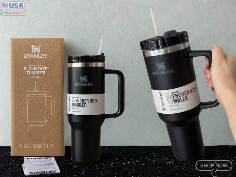May include: Two black Stanley Flowstate Quencher H2.0 tumblers with handles, straws, and black lids. One tumbler stands next to its brown cardboard packaging, while the other is held. The packaging features the Stanley logo and product name.