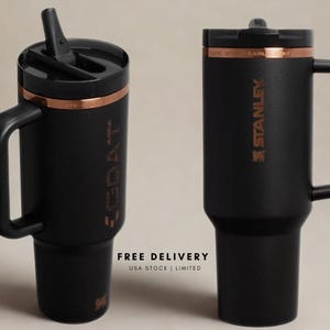 May include: Two matte black tumblers with handles and black lids. One tumbler features a straw and the text GOAT vertically, while the other displays the Stanley logo. Both have a copper-coloured band near the top rim and a tapered base design.