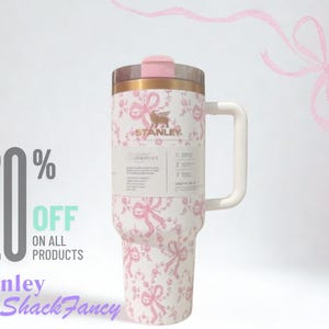 K&ouml;nnte beinhalten: Ein wei&szlig;er Stanley-Becher mit rosa Schleifen- und Blumenmuster, ausgestattet mit einem goldenen Rand, einem rosa Deckel und einem wei&szlig;en Henkel. Ein Produktetikett ist vorne angebracht. Der Text lautet 20% OFF ON ALL PRODUCTS und Stanley LoveShackFancy.