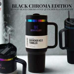 May include: Three matte black Stanley Quencher H2.0 Flowstate tumblers with iridescent rainbow-colored rims. The central tumbler features a white paper band with the text 'QUENCHER H2.0 TUMBLER' and a black handle. One tumbler shows steam rising.