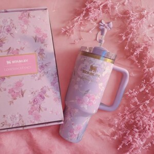 May include: A lavender Stanley tumbler with a floral pattern and a matching box featuring the text Stanley and LoveShackFancy. The tumbler includes a handle, a clear lid, and a straw topper shaped like a bow. The floral design features pink roses.