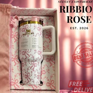 Puede incluir: Un vaso Stanley blanco con un patr&oacute;n de lazos y flores rosas, con un logotipo dorado, una tapa rosa claro y un asa color crema. El vaso se presenta en una caja a juego junto a una bolsa de tela rosa, con el texto que dice RIBBION ROSE.
