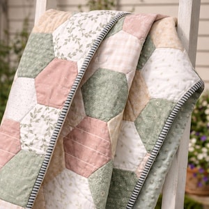 Puede incluir: Colcha hecha a mano con diseño de patchwork. La colcha presenta parches hexagonales en tonos rosa pálido, verde salvia y crema. Algunos parches tienen estampados florales, otros son lisos o a rayas. La colcha tiene un borde a rayas.