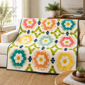 Op de afbeelding: Een kleurrijke quilt met een bloemenpatroon in roze, oranje, geel en teal tinten. De quilt is over een witte bank gedrapeerd en toont het ingewikkelde ontwerp en de levendige kleuren. De randen van de quilt zijn afgewerkt met een donkerblauwe bies.