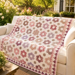 May include: A floral quilt in shades of pink, purple, and brown, draped over a white sofa. The quilt features a repeating pattern of flower-like shapes and geometric accents. The quilt's border has a triangle pattern.