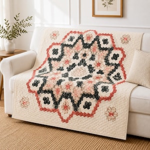 May include: A cream-colored sofa cover with a large, central floral design in shades of pink, black, and red. The cover is quilted and features a geometric pattern. Additional floral motifs are placed around the edges.