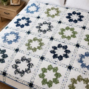 Può includere: Una trapunta bianca con un motivo ripetuto di disegni a forma di stella nei toni del blu e del verde. La trapunta è divisa in quadrati con un bordo a motivi, creando un aspetto geometrico e strutturato. La biancheria da letto è probabilmente in cotone.