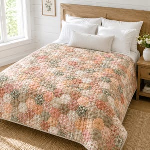 Op de afbeelding: Een bed is bedekt met een bloemenquilt in roze, perzik en groene tinten. De quilt heeft een zeshoekig patroon en wordt aangevuld met witte kussens en een licht kussen. Het bed staat tegen een houten hoofdbord.