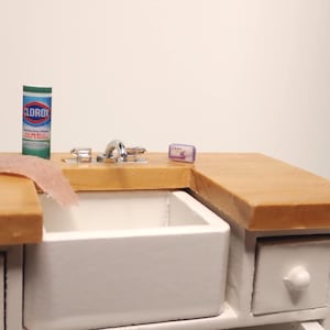 May include: Miniature kitchen scene featuring a white sink, wooden countertop, and a small white cabinet. A Clorox disinfecting wipes container, a pink sponge, and a bar of Dove soap are on the countertop. The faucet is chrome.