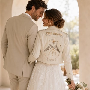 May include: A cream-colored leather jacket featuring an embroidered design on the back with the text TILL DEATH, depicting two skeleton hands reaching toward each other with a star, lightning bolts, and clouds in a circular frame.