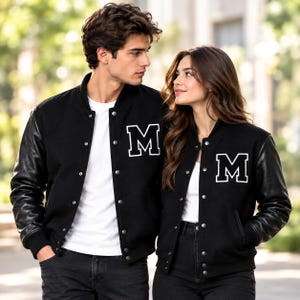 Könnte beinhalten: Eine schwarze Collegejacke mit langen schwarzen Ärmeln, einem weißen Buchstaben-Aufnäher M auf der Brust und einer Knopfleiste vorne. Die Jacke wird über einem schlichten weißen T-Shirt getragen und mit einer dunklen Jeans kombiniert.