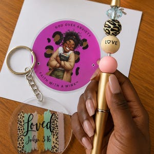 May include: A gold-toned pen with decorative beads including a leopard print sphere, a light pink sphere, and a sphere labelled LOVE. Beside it is a circular keychain with a leopard print background, a cartoon figure, and the text GOD OVER ANXIETY FAITH WITH A WINK, plus a clear acrylic keychain with the text loved John 3:16.