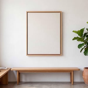 May include: A rectangular canvas art piece featuring a blank, off-white fabric surface encased in a light-toned wooden frame. The artwork is mounted on a plain wall above a long, simple wooden bench, positioned near a potted green plant.