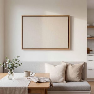 Puede incluir: Un espacio de vida de tonos neutros con una gran obra de arte en lienzo enmarcada sobre un banco gris con almohadas. Una mesa de madera está decorada con un jarrón con vegetación, una taza y un camino de mesa de lino. El ambiente es minimalista y moderno.