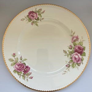 May include: A round cream-coloured ceramic dinner plate featuring a delicate floral pattern of pink roses and green leaves arranged in three clusters. The plate has a scalloped edge finished with a thin gold trim along the entire outer rim.