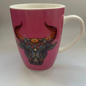 Peut inclure: Un mug en céramique rose avec une illustration détaillée et colorée d'une tête de taureau sur le devant. Le motif du taureau intègre des motifs tourbillonnants complexes dans des tons orange, bleu et jaune. Le mug a un intérieur et une anse blancs.