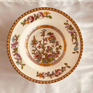 May include: A round ceramic bowl with an off-white base color, featuring a central floral motif with green, pink, and yellow tones. The rim is decorated with a brown geometric Greek key pattern and additional floral sprays in pink and blue.