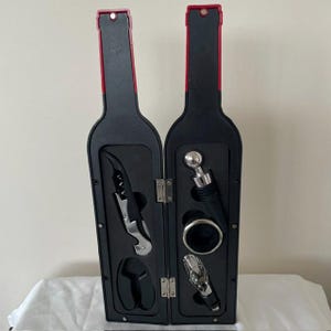 May include: A black wine bottle-shaped case that opens to reveal a wine accessory set. The set includes a silver and black corkscrew, a silver bottle stopper with a round top, a silver drip ring, and a silver pourer, all held in foam inserts.