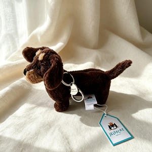 Könnte beinhalten: Ein Dackel-Schlüsselanhänger aus dunkelbraunem Plüsch mit hellbrauner Schnauze und Ohren. Das Stofftier hat einen silbernen Metallring am Rücken und ein weißes Papieretikett mit dem Jellycat-Logo, das eine schwarze Katze mit orangefarbenem Hut zeigt.