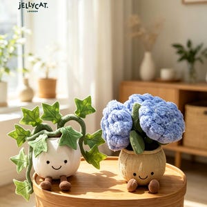 May include: Two plush plant toys sit on a wooden table. One is a white pot with green ivy, the other is a tan pot with blue flowers. Both have smiling faces and small brown feet. The Jellycat London logo is in the top left corner.