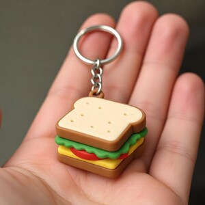 Peut inclure: Un porte-clés en forme de sandwich. Le porte-clés représente un sandwich miniature avec des couches de pain, de laitue, de tomate et de fromage. Le porte-clés est attaché à un anneau en métal argenté.