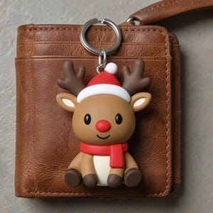 以下が含まれることがあります： 赤い鼻、スカーフ、サンタ帽をかぶったクリスマスをテーマにしたトナカイのキーホルダー。トナカイは茶色、白、赤で、シルバーのキーリングが付いています。キーホルダーは茶色の革の財布の上にあります。