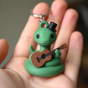 Pode incluir: Um chaveiro de cobra verde com uma cartola preta e um ukulele castanho. A cobra está enrolada e tem olhos pretos. O chaveiro é segurado em uma mão.