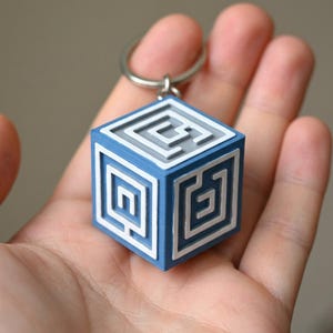 May include: A blue and white cube-shaped keychain featuring a raised maze pattern on its visible faces. The object is constructed with a matte finish and includes a silver-toned metal split ring attached to the top corner for secure key storage.