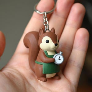 Könnte beinhalten: Ein brauner Eichhörnchen-Schlüsselanhänger mit cremefarbenem Gesicht und Bauch, der eine dunkelgrüne Schürze trägt. Das Eichhörnchen hält eine kleine, runde Stoppuhr mit weißem Zifferblatt, schwarzem Zeiger und dem Buchstaben L, befestigt an einem Metallring.