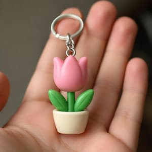 Op de afbeelding: Een roze tulp sleutelhanger met groene bladeren en een groene stengel, in een crème pot. De sleutelhanger heeft een zilveren ring. Het ontwerp is een miniatuur, cartoonachtige weergave van een tulp in een pot.