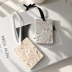 May include: Two rectangular hanging ornaments featuring a raised ginkgo leaf pattern. One ornament is cream-coloured with a white ribbon, and the other is light grey with a black ribbon. Both pieces have a textured surface and a hole for hanging.