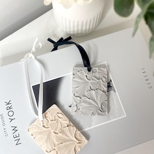 May include: Two rectangular, textured ornaments with ginkgo leaf patterns. One is a light beige colour, the other is grey. Each has a ribbon for hanging. The ornaments rest on a book with the text "NEW YORK CITY GUIDE".