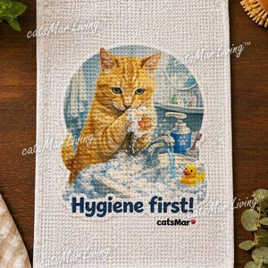 Può includere: Un canovaccio bianco con trama a nido d'ape che presenta una stampa di un gatto tigrato arancione che si lava le zampe in un lavandino con bolle di sapone, un dispenser di sapone blu e una papera di gomma gialla, con il testo Hygiene first! catsMar.