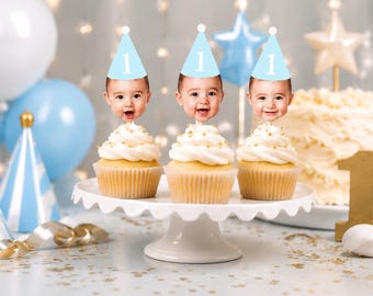 May include: Three cupcakes with white frosting and gold sprinkles, each topped with a cutout of a baby's face wearing a light blue party hat featuring the number 1. The cupcakes are arranged on a white scalloped cake stand.
