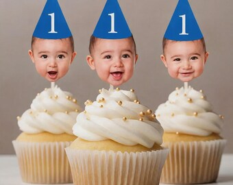May include: Three cupcakes with white frosting and gold sprinkles. Each cupcake is topped with a blue party hat with the number 1 and a photo of a baby's face. The cupcakes are in white paper liners.