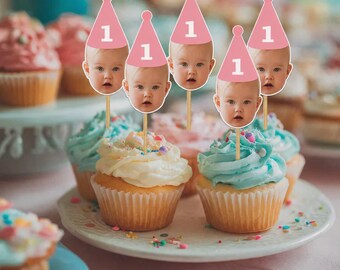 May include: Cupcakes with light blue and white frosting, topped with colorful sprinkles. Pink party hat toppers with a baby's face and the number 1 are inserted into the cupcakes, perfect for a first birthday celebration.
