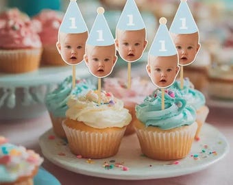 May include: Five cupcake toppers featuring a photographic print of a baby's face wearing a light blue party hat with the number 1 printed in white. Each topper is attached to a thin wooden stick, designed to be inserted into frosted cupcakes.