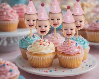 May include: A collection of cupcakes with colorful frosting and sprinkles, decorated with baby face toppers wearing pink party hats with the number 1. The cupcakes are arranged on a white plate, perfect for a first birthday celebration.