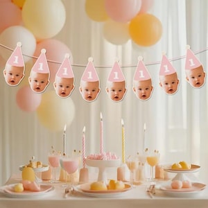 May include: A first birthday party setting with pink and yellow balloons, a banner of baby faces wearing party hats with the number 1, and a table set with candles, desserts, and drinks. The theme is a celebration.