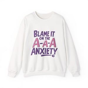 May include: White sweatshirt with the text "Blame it on the A-A-A Anxiety" in purple and pink lettering. The sweatshirt has a crew neck and long sleeves. The design is centered on the front of the garment.