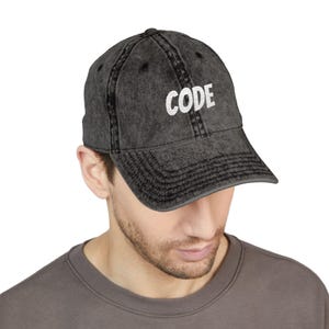 May include: A dark gray baseball cap with the word "CODE" in white block letters. The cap has a distressed, vintage look, with a curved brim and adjustable strap. The hat is made of a soft, worn material.